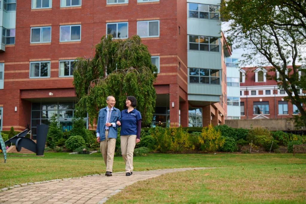 Two people walk arm-in-arm on a path near a red brick building, surrounded by greenery.