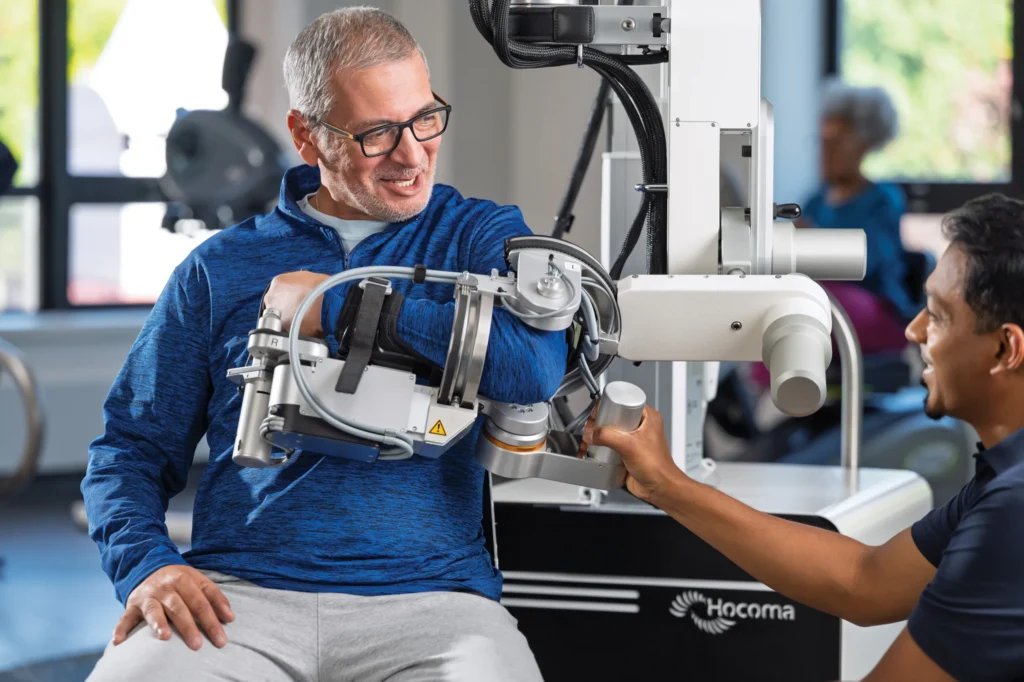 Man uses robotic arm rehabilitation device with therapist assistance in a clinic setting.