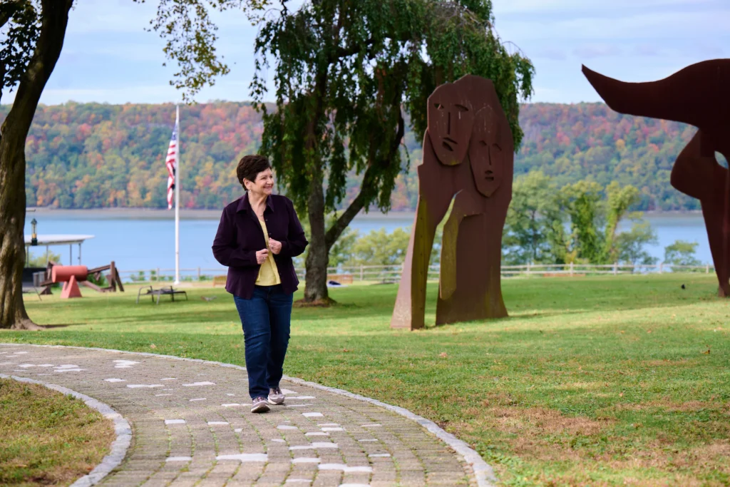 Woman walks on park path by large metal sculptures, lake, and autumn trees in background.