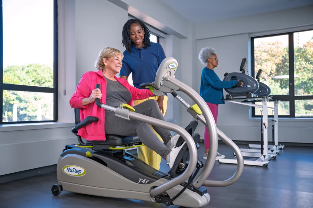 Three women exercising indoors: one on a NuStep with trainer assistance, another walking on treadmill.