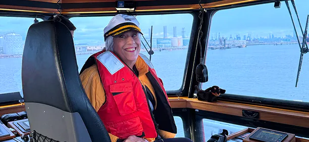 Smiling person in captain's hat and red life vest steering a boat on the water.