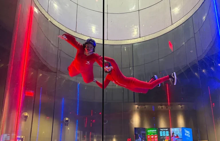 Two people in red suits float mid-air inside a wind tunnel with blue and red lighting.