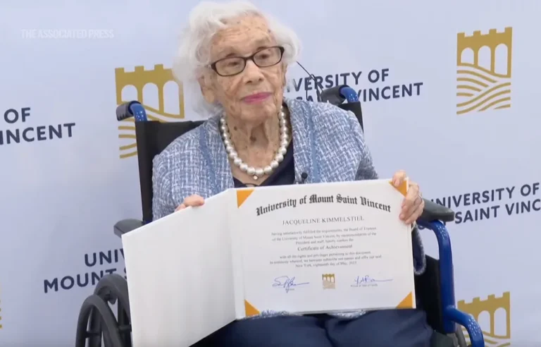 Elderly woman in wheelchair smiles, holding certificate before University of Saint Vincent banner.