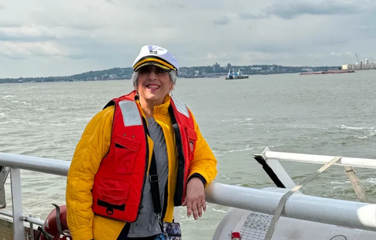 Smiling person in captain’s hat and life jacket stands on a boat with water and land behind.