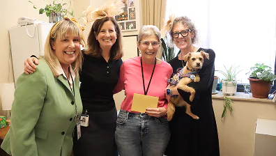 Four women, one holding a small dog, standing in an office with plants and photos behind them.