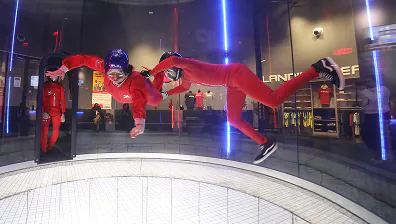Two people in red suits and helmets indoor skydiving in a blue-lit vertical wind tunnel.