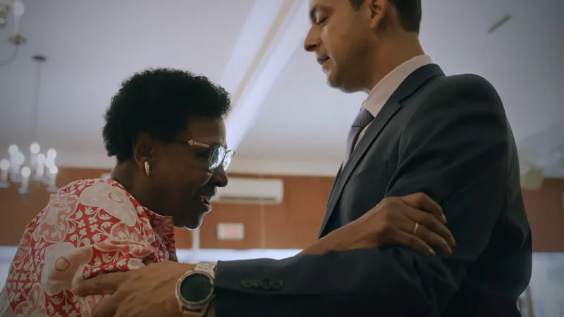Elderly woman smiling and holding hands with a man in a suit in a well-lit room.