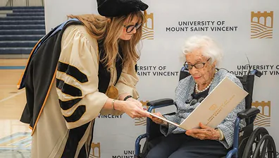 Woman in academic regalia gives diploma to elderly woman in wheelchair at ceremony.