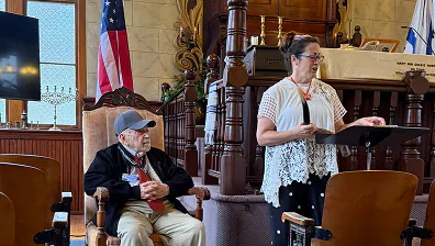 Woman speaks at podium; elderly man sits nearby in room with flags and wooden furniture.