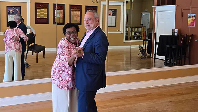 Two people smile and dance together in a studio with wood floors and a mirrored wall.
