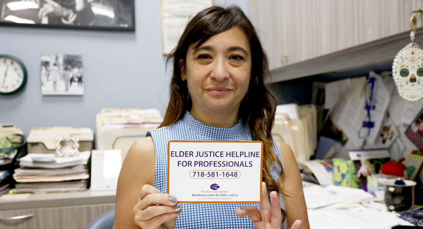 Woman in office holds sign: “Elder Justice Helpline for Professionals 718-581-1648”.