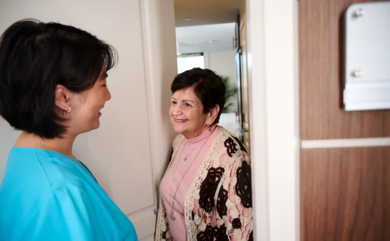 A RiverSpring Living care aide in scrubs smiles while talking with an older woman at the doorway of her Riverdale apartment
