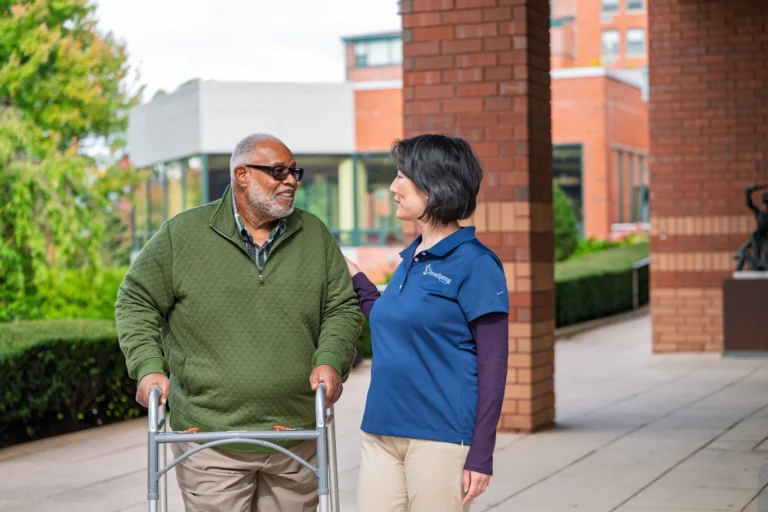 Older man with walker talks to woman in blue shirt outside a brick building.