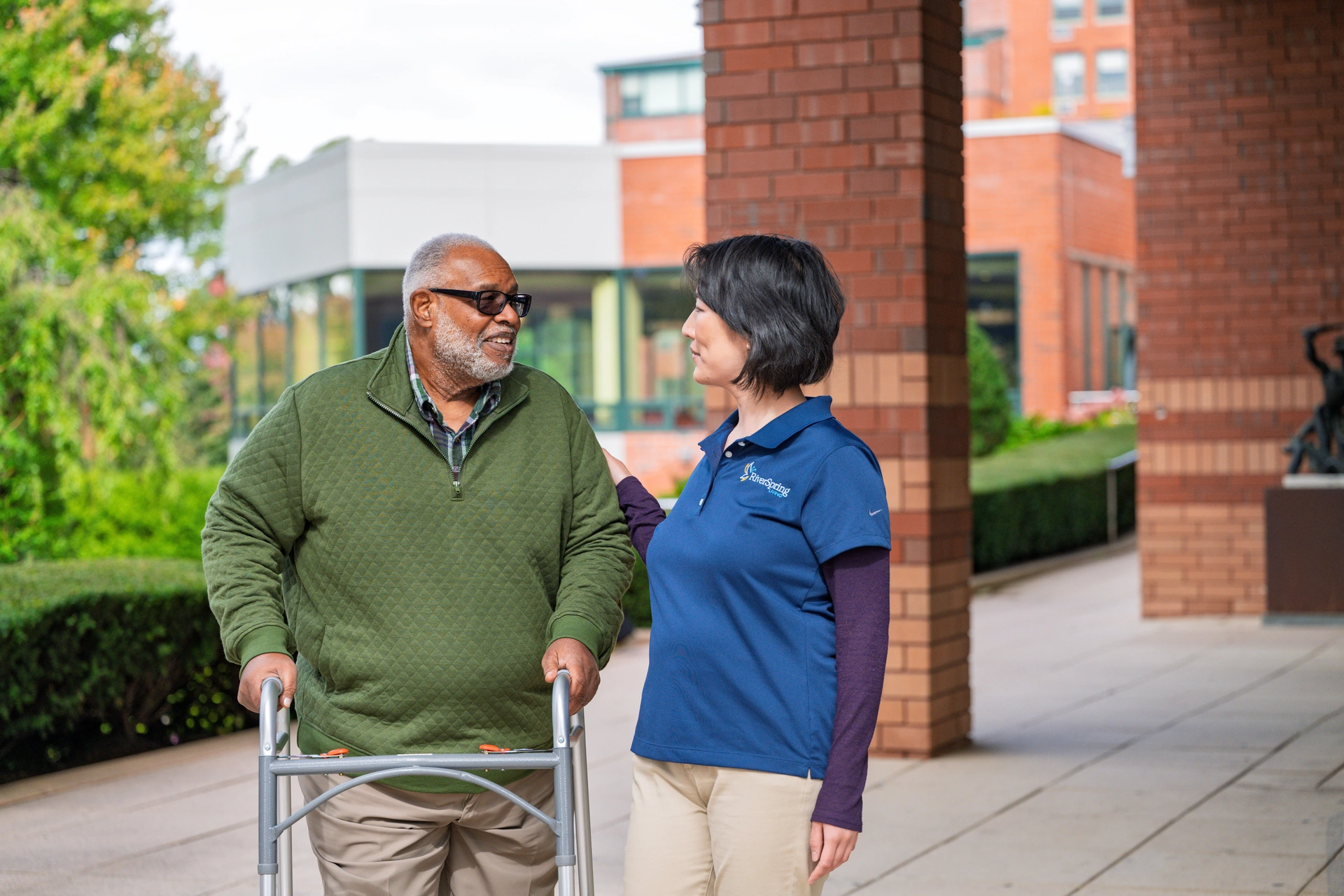 Older man with walker talks to woman in blue shirt outside a brick building.